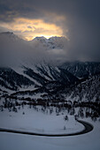 Sonnenuntergang durch die Wolken über der Cima Di Saoseo und der verschneiten Strasse zum Berninapass, Val Poschiavo, Kanton Graubünden, Schweiz
