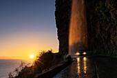 Auto mit eingeschalteten Scheinwerfern fährt in der Abenddämmerung unter dem Anjos-Wasserfall hindurch, Ponta do Sol, Insel Madeira, Portugal