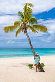 Schöne Frau bewundert das türkisfarbene Meer an eine Palme gelehnt an einem weißen Sandstrand, Barbuda, Antigua und Barbuda, Karibik