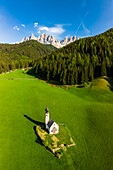 Iconic church of Ranui and Odle group surrounded by green woods in spring, overhead view, Funes Valley, South Tyrol, Italy