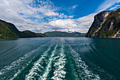 Das Kielwasser eines Schiffes zieht durch den Geirangerfjord, der von schroffen, steilen Bergen gesäumt wird. Geirangerfjord, Norwegen.