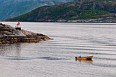 A small motor boat crosses the mountain lined glacial waters of Svesfjorden near a small lighthouse. Svesfjorden, Norway.