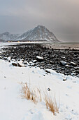 A snow-covered coastline with a rocky beach, mountains, and a village. Noss, Vesteralen Islands, Nordland, Norway.