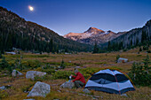 Backpacking camper in Lostine River valley meadow with moon rising over Eagle Cap mountain; Eagle Cap Wilderness, Wallowa Mountains, Oregon.