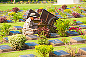 Männlicher Gärtner pflegt Gräber auf dem Kriegsfriedhof Kanchanaburi, Thailand, Südostasien, Asien