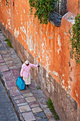 Elderly woman walking along street with her hand on a wall to steady herself; San Miguel de Allende, Guanajuato, Mexico.