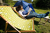 Camperin sitzt auf einem Liegestuhl und liest, mit Zelt im Hintergrund