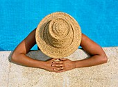 Blick von oben auf eine Frau mit Strohhut, die sich an den Rand eines Swimmingpools lehnt