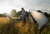 Frau schüttelt Schlafsack neben Zelt in Wiese aus, Sonnenuntergang