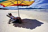Man Sitting On Deckchair Under Umbrella On Beach, Side View