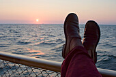 Füße einer Person auf einem Geländer am See bei Sonnenuntergang, See Vanern, Schweden