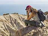 Eine junge Frau sitzt auf einem Felsen, blickt zurück und posiert für die Kamera mit Blick auf das Naturreservat Ein Gedi, Bezirk Totes Meer; Region Süd, Israel.