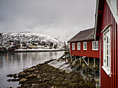 A Red Building Along The Water's Edge With Snow Covered Hills Under A Cloudy Sky; Lofoten Islands, Nordland, Norway