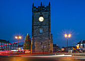 Die Uhr in einem Turm und die Laternenpfähle leuchten in der Dämmerung, mit Lichtspuren auf der Straße; Richmond, Yorkshire, England