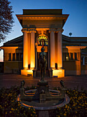 A building illuminated with golden light at dusk with a clear sky and a water fountain in a garden; Belgrade, Vojvodina, Serbia