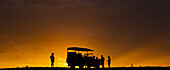 Silhouette von Touristen auf einer Luxussafari, die neben einem Freizeitfahrzeug in der Wüste bei Sonnenuntergang stehen; Sossusvlei, Hardap Region, Namibia.