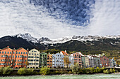 Bunte Gebäude entlang des Flussufers mit schneebedeckten Berggipfeln, dramatischen Wolken und blauem Himmel über dem Wasser; Innsbruck, Tirol, Österreich.