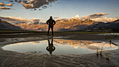 A man stands with his reflection in a pool of water looking out over the Saint Elias Mountains at sunset, Kluane National Park and Reserve; Destruction Bay, Yukon, Canada
