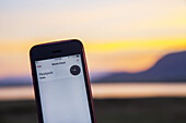 Ein Handy zeigt die Ortszeit in Reykjavik kurz vor Mitternacht an, wobei das Tageslicht zur Sommersonnenwende noch sichtbar ist, wenn die Sonne in Island selbst um Mitternacht noch nicht vollständig untergeht; Reykjavik, Island