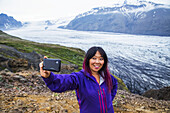 Eine asiatische Wanderin macht ein Selbstporträt auf dem Berggipfel mit dem Gletscher im Hintergrund im Vatnajokull-Nationalpark; Island