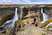 Eine junge asiatische Wanderin posiert für ein Porträt am Rande einer atemberaubenden Landschaft mit zwei Wasserfällen, bekannt als Haifoss; Island
