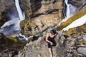 Eine junge asiatische Wanderin posiert für ein Porträt am Rande einer atemberaubenden Landschaft mit zwei Wasserfällen, die als Haifoss bekannt ist; Island