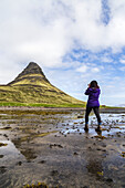 Eine Wanderin bleibt bei Ebbe am Strand stehen, um ein Foto mit einer Spiegelreflexkamera vor dem Berg Kirkjufell auf der Halbinsel Snaefellsnes im Westen Islands zu machen; Island