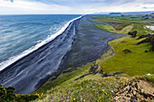 The view from the top of the tourist lookout at Dyrholaey near Vik, Southern Iceland. The black sand beach extends for miles into the distance and green pasture and farmland is seen in the background; Vik, Iceland