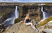 Eine junge Wanderin posiert für ein Porträt am Rande eines atemberaubenden Wasserfalltals, bekannt als Haifoss, in Südisland; Island