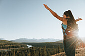 Eine Frau, die mit ausgestreckten Armen auf einem Wanderweg in den Rocky Mountains in der Nähe von Hinton, Alberta, Kanada, steht und von Sonnenstrahlen angestrahlt wird.