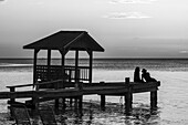 A couple sits on the end of a dock looking out to the tranquil ocean at sunset; Roatan, Bay Islands Department, Honduras