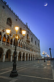 Dogenpalast in der Abenddämmerung mit beleuchteten Laternenpfählen und einem Mond am blauen Himmel; Venedig, Italien.