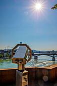 Binoculars along the waterfront looking out to the Rhine River with bridge and a sunburst in the blue sky; Basel, Basel Stadt, Switzerland
