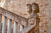Carved stone sculpture of human heads and an animal face as a decorative post on a handrail beside a brick wall and stairway; Venice, Italy