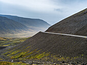 Road going around a bend on a hillside with a view of a valley and mountains under an overcast sky; Westfjords, Iceland