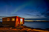 A small house on the water's edge with a view of the tranquil coastline at nightfall and the glowing northern lights above; Nuuk, Sermersooq, Greenland