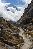 Gokyo trek with autumn colored tundra and rock cairns lining the trail, while  clouds partially obscure the snow capped Himalayan mountain peak in the background on a sunny day near Gokyo, Sagarmatha National Park; Solokhumbu district, Nepal