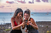 Two young women looking at digital photos of the sunset over Airlie Beach Harbor, on their smart phone; Airlie Beach, Whitsunday Region, Queensland, Australia