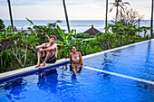 Man and woman sitting poolside at resort, overlooking surfing beach of Balian Beach; Selemadeg Barat, Tabanan, Bali, Indonesia