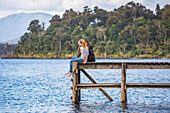 Three friends sitting on the edge of a wooden dock at Lake Mahinapua; West Coast, New Zealand