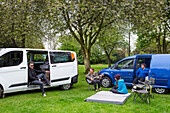 Friends relaxing while setting up camp outside two modified caravan campers; Bourton-on-the-Water, England, United Kingdom