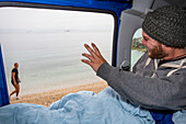 Free camping and waking up on the coastal beaches of Split, Croatia. A man looks at the ocean from the bed inside of his vehicle, and waves to a woman passing by; Split, Splitsko-dalmatinska zupanija, Croatia