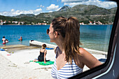 Eine Gruppe hält in Slano für einen Nachmittag am Strand. Eine junge Frau im Badeanzug und mit Sonnenbrille steht am Fahrzeug und schaut auf den Strand und das Wasser; Slano, Dubrovacko-neretvanska zupanija, Kroatien