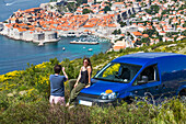 Travellers explore the surrounding cliffs above Dubrovnik with their vehicle to catch the stunning views over the coastal city. A young couple takes photographs by their camper van; Dubrovnik, Dubrovacko-neretvanska zupanija, Croatia
