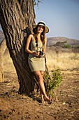 Porträt einer Frau, die an einem Baumstamm lehnt, ein Sonnenkleid und einen Strohhut trägt, eine Kamera hält und in die Kamera lächelt, an einem sonnigen Tag auf der Gabus Game Ranch; Otavi, Otjozondjupa, Namibia.