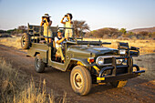 Frauen in einem Jeep, eine mit einem Fernglas und eine mit einer Kamera, schauen in die Savanne, während der Fahrer hinter dem Lenkrad sitzt, während einer Safari auf der Gabus Game Ranch; Otavi, Otjozondjupa, Namibia.
