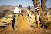 Safari guide explaining termite mound bloodstain to women travelers on the savanna at the Gabus Game Ranch at sunset; Otavi, Otjozondjupa, Namibia