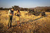 Safari guide showing ground sample to woman traveler holding a camera on the savanna with another woman standing in a jeep looking at a reference book in the background at the Gabus Game Ranch; Otavi, Otjozondjupa, Namibia