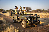 Safari guide and woman traveler standing in a parked jeep looking at reference book while another kneels in jeep looking out onto the savanna with binoculars at the Gabus Game Ranch; Otavi, Otjozondjupa, Namibia
