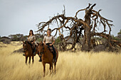 Zwei Frauen auf Pferden (Equus ferus caballus), die in die Kamera lächeln und in der Nähe eines toten Baumes im Gebüsch der Savanne auf der Gabus Game Ranch stehen; Otavi, Otjozondjupa, Namibia.